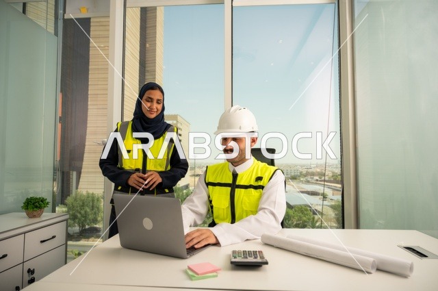 Collaboration to complete work quickly and accurately, auditing and supervising engineering projects from an engineering office. Two Arab Gulf engineers from Saudi Arabia wearing safety helmets and work vests are studying and evaluating engineering plans using a laptop, integrating technology with work.