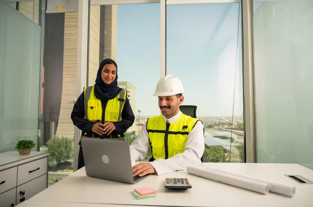 Collaboration to complete work quickly and accurately, auditing and supervising engineering projects from an engineering office. Two Arab Gulf engineers from Saudi Arabia wearing safety helmets and work vests are studying and evaluating engineering plans using a laptop, integrating technology with work.