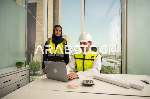 الإشراف والمناقشة على المشاريع الهندسية من مكتب هندسي، دمج التكنلوجيا مع العمل، التعاون لإنجاز العمل بسرعة ودقة, مهندسان عربيان خليجيان سعوديان يرتديان خوذة الحماية وسترة العمل و يقومان بدراسة و تقييم الخطط الهندسية من خلال الحاسوب المحمول