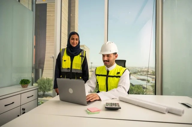 الإشراف والمناقشة على المشاريع الهندسية من مكتب هندسي، دمج التكنلوجيا مع العمل، التعاون لإنجاز العمل بسرعة ودقة, مهندسان عربيان خليجيان سعوديان يرتديان خوذة الحماية وسترة العمل و يقومان بدراسة و تقييم الخطط الهندسية من خلال الحاسوب المحمول