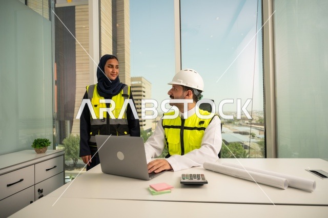 The concept of construction and architecture, supervising and discussing engineering projects from an engineering office, collaborating to complete work quickly and accurately. Two Arab Gulf engineers from Saudi Arabia are wearing work vests and are studying and evaluating engineering plans using a laptop.