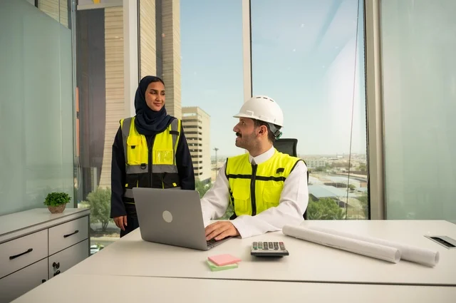 مفهوم الإنشاءات والهندسة المعمارية، الإشراف والمناقشة على المشاريع الهندسية من مكتب هندسي، التعاون لإنجاز العمل بسرعة ودقة, مهندسان عربيان خليجيان سعوديان يرتديان سترة العمل و يقومان بدراسة و تقييم الخطط الهندسية من خلال الحاسوب المحمول