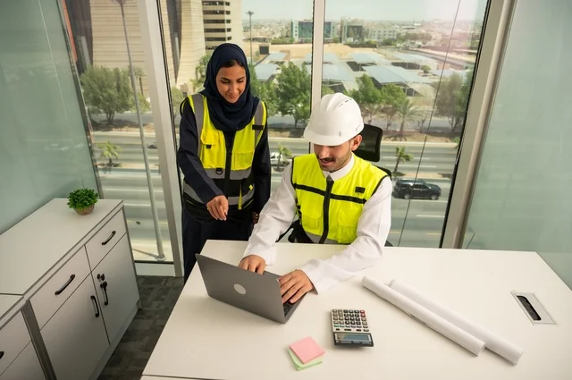 Integrating technology with work, collaborating to complete tasks quickly and accurately, auditing and supervising engineering projects from an engineering office. Two Arab Gulf engineers from Saudi Arabia are wearing safety helmets and work vests and are studying and evaluating engineering plans using a laptop.