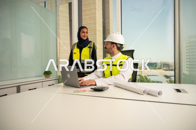 الإشراف والمناقشة على المشاريع الهندسية من مكتب هندسي، دمج التكنلوجيا مع العمل، التعاون لإنجاز العمل بسرعة ودقة, مهندسان عربيان خليجيان سعوديان يرتديان خوذة الحماية وسترة العمل و يقومان بدراسة و تقييم الخطط الهندسية من خلال الحاسوب المحمول