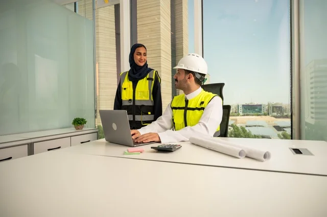 الإشراف والمناقشة على المشاريع الهندسية من مكتب هندسي، دمج التكنلوجيا مع العمل، التعاون لإنجاز العمل بسرعة ودقة, مهندسان عربيان خليجيان سعوديان يرتديان خوذة الحماية وسترة العمل و يقومان بدراسة و تقييم الخطط الهندسية من خلال الحاسوب المحمول