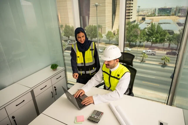 التعاون لإنجاز العمل بسرعة ودقة، التدقيق والإشراف على المشاريع الهندسية من مكتب هندسي, مهندسان عربيان خليجيان سعوديان يرتديان خوذة الحماية وسترة العمل و يقومان بدراسة و تقييم الخططت الهندسية من خلال الحاسوب المحمول، دمج التكنلوجيا مع العمل