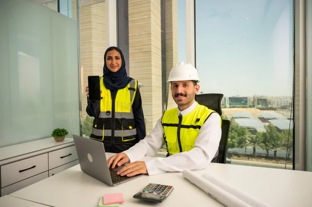 مهندسان عربيان خليجيان سعوديان يرتديان سترة العمل و يقومان بالعمل من خلال الحاسوب المحمول، تعرض المهندسة خلفية هاتف محمول سوداء، دمج التكنولوجيا والأجهزة التقنية في الحياة العملية، التعاون لإنجاز العمل بسرعة ودقة, الإشراف والمناقشة على المشاريع الهندسية