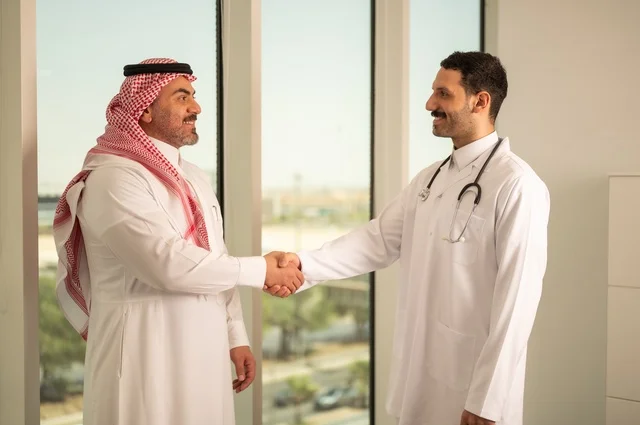 المصافحة والقاء التحية والسلام، مفهوم الفحص الطبي، العمل في القطاع الصحي، تقديم الخدمات العلاجية للمرضى, طبيب عربي خليجي سعودي يرتدي الثوب التقليدي والمعطف الطبي يضع السماعة الطبية يقوم بمصافحة مريضه العمل في المراكز الصحية والمستشفيات