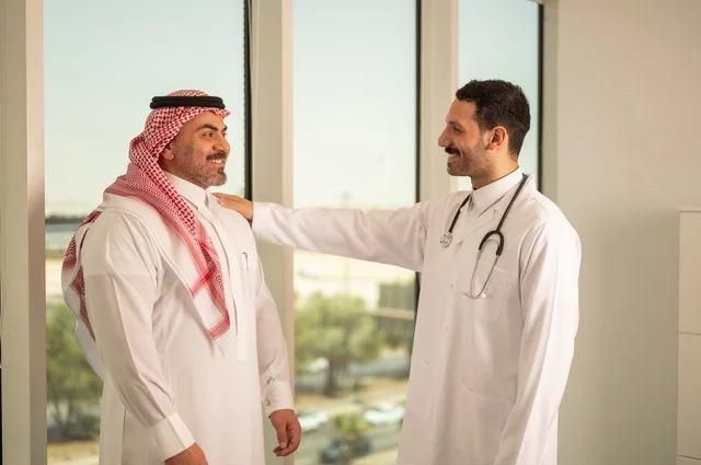 مفهوم الفحص الطبي، العمل في القطاع الصحي، تقديم الخدمات العلاجية للمرضى, طبيب عربي خليجي سعودي يرتدي الثوب التقليدي والمعطف الطبي يضع السماعة الطبية يقف مع مريضه ويعمل في المراكز الصحية والمستشفيات