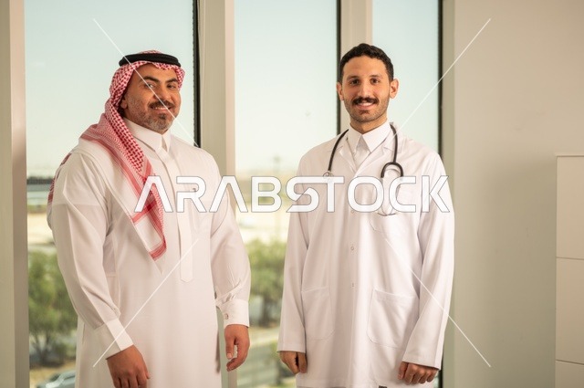 مفهوم الفحص الطبي، العمل في القطاع الصحي، تقديم الخدمات العلاجية للمرضى, طبيب عربي خليجي سعودي يرتدي الثوب التقليدي والمعطف الطبي يضع السماعة الطبية يقف مع مريضه ويعمل في المراكز الصحية والمستشفيات