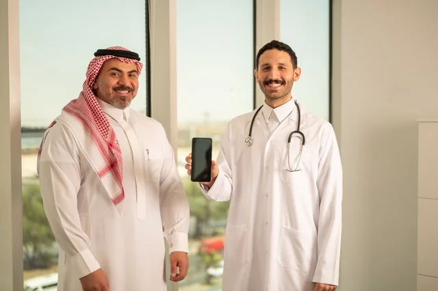 عرض شاشة سوداء فارغة عبر الهاتف المحمول، مفهوم الفحص الطبي، العمل في القطاع الصحي، تقديم الخدمات العلاجية للمرضى, طبيب عربي خليجي سعودي يرتدي الثوب التقليدي والمعطف الطبي يضع السماعة الطبية يقف مع مريضه ويعمل في المراكز الصحية والمستشفيات