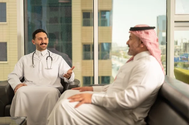 مفهوم الفحص الطبي، العمل في القطاع الصحي، تقديم الخدمات العلاجية للمرضى, طبيب عربي خليجي سعودي يرتدي الثوب التقليدي والمعطف الطبي يضع السماعة الطبية يجلس مع مريضه ويعمل في المراكز الصحية والمستشفيات