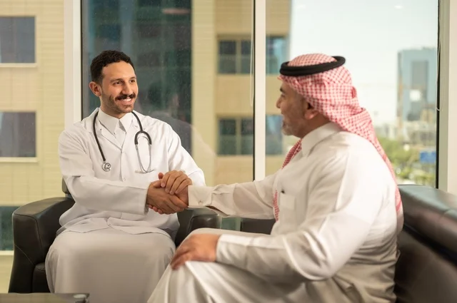 المصافحة والقاء التحية والسلام، مفهوم الفحص الطبي، العمل في القطاع الصحي، تقديم الخدمات العلاجية للمرضى, طبيب عربي خليجي سعودي يرتدي الثوب التقليدي والمعطف الطبي يضع السماعة الطبية يقوم بمصافحة مريضه العمل في المراكز الصحية والمستشفيات