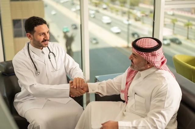 المصافحة والقاء التحية والسلام، مفهوم الفحص الطبي، العمل في القطاع الصحي، تقديم الخدمات العلاجية للمرضى, طبيب عربي خليجي سعودي يرتدي الثوب التقليدي والمعطف الطبي يضع السماعة الطبية يقوم بمصافحة مريضه العمل في المراكز الصحية والمستشفيات