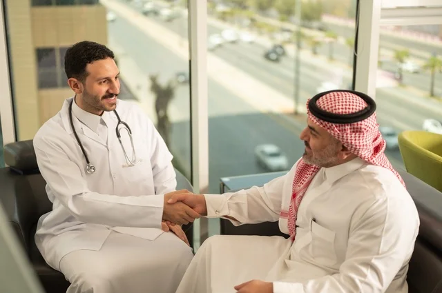 المصافحة والقاء التحية والسلام، مفهوم الفحص الطبي، العمل في القطاع الصحي، تقديم الخدمات العلاجية للمرضى, طبيب عربي خليجي سعودي يرتدي الثوب التقليدي والمعطف الطبي يضع السماعة الطبية يقوم بمصافحة مريضه العمل في المراكز الصحية والمستشفيات