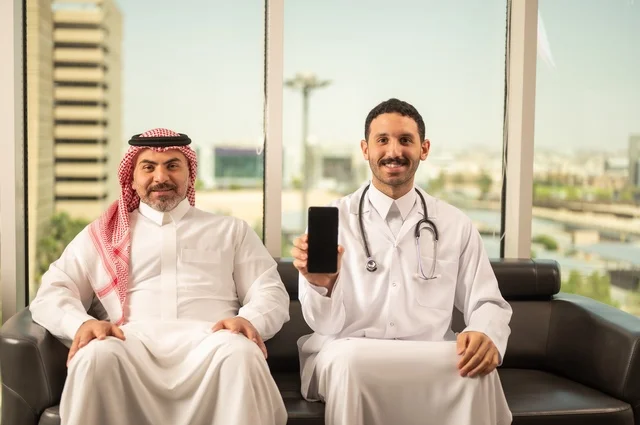 عرض شاشة سوداء فارغة عبر الهاتف المحمول، مفهوم الفحص الطبي، العمل في القطاع الصحي، تقديم الخدمات العلاجية للمرضى, طبيب عربي خليجي سعودي يرتدي الثوب التقليدي والمعطف الطبي يضع السماعة الطبية يجلس مع مريضه ويعمل في المراكز الصحية والمستشفيات