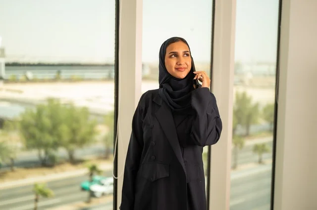 إجراء اتصال هاتفي عبر الجوال, استخدام جهاز تقني حديث ومتطور في العمل, مهنة ووظيفة مكتبية في السعودية, امرأة عربية خليجية سعودية محجبة ترتدي العباءة تقف في المكتب وتمسك الهاتف المحمول بيدها,التواصل مع العملاء عن بعد