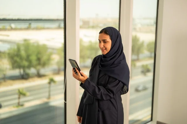 إجراء اتصال هاتفي عبر الجوال, استخدام جهاز تقني حديث ومتطور في العمل, مهنة ووظيفة مكتبية في السعودية, امرأة عربية خليجية سعودية محجبة ترتدي العباءة تقف في المكتب وتمسك الهاتف المحمول بيدها,التواصل مع العملاء عن بعد