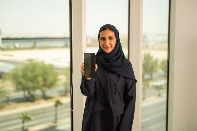عرض شاشة سوداء فارغة عبر الجوال, استخدام جهاز تقني حديث ومتطور في العمل, مهنة ووظيفة مكتبية في السعودية, امرأة عربية خليجية سعودية محجبة ترتدي العباءة تقف في المكتب وتمسك الهاتف المحمول بيدها, النظر الى الكاميرا بايماءات السرور