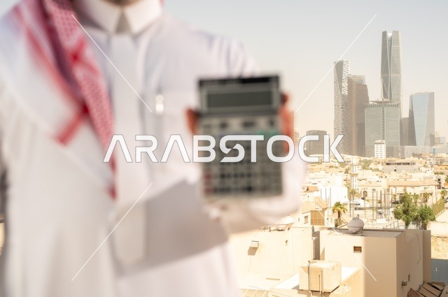مفهوم تسهيل الاعمال والقدرة على إنجاز المهام، استخدام التقنيات الحديثة والأجهزة المتطورة ، رجل عربي خليجي سعودي يرتدي الثوب التقليدي يمسك بيده آلة حاسبة لإجراء عمليات الحساب، خلفية الابراج وناطحات السحاب في الرياض