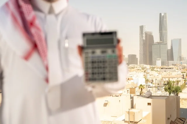 The concept of facilitating business and the ability to accomplish tasks, using modern technologies and advanced devices, a Saudi Arabian Gulf man wearing traditional clothing holding a calculator to perform calculations, a background of towers and skyscrapers in Riyadh