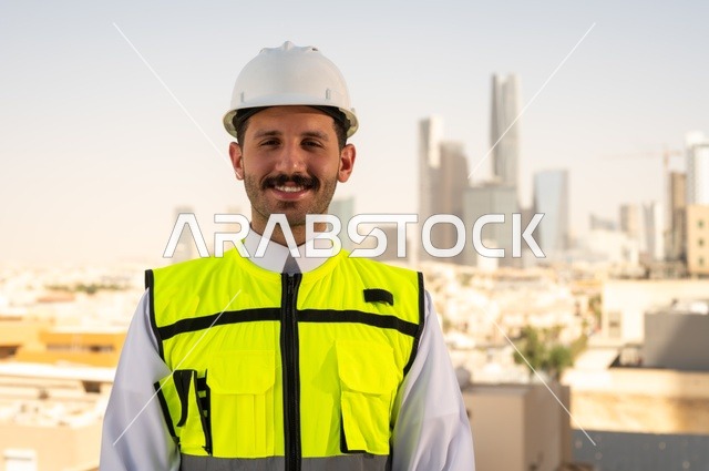 الاهتمام بالتطور العمراني بسواعد المهندسين السعوديين, مهندس عربي خليجي سعودي يرتدي سترة العمل وخوذة الحماية البيضاء ينجز المهام اليومية في المكتب، مفهوم الهندسة المعمارية، مشاريع إعمارية سعودية وطنية، خلفية أبراج الرياض