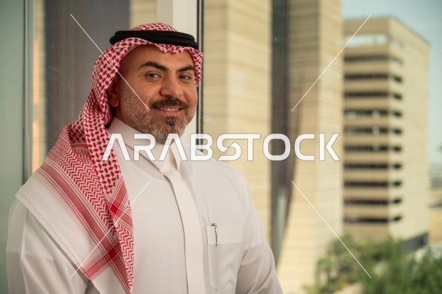 الاناقة والاهتمام بالمظهر الخارجي. صورة مقربة لرجل عربي خليجي سعودي مبتسم يرتدي الثوب والشماغ التقليدي ينظر الى الكاميرا بايماءات السرور والسعادة. الخروج في الهواء الطلق،  مفهوم الرجولة والثقة بالنفس، خلفية أبنية