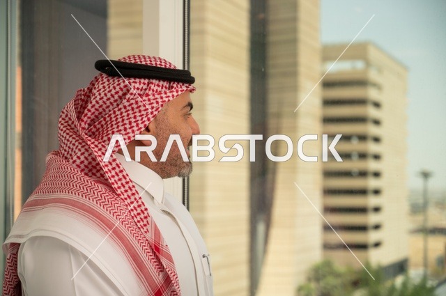 مفهوم الرجولة والثقة بالنفس، الاناقة والاهتمام بالمظهر الخارجي. صورة جانبية مقربة لرجل عربي خليجي سعودي مبتسم يرتدي الثوب والشماغ التقليدي ينظر الى الكاميرا بايماءات السرور والسعادة. الخروج في الهواء الطلق،   خلفية أبنية