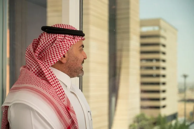 مفهوم الرجولة والثقة بالنفس، الاناقة والاهتمام بالمظهر الخارجي. صورة جانبية مقربة لرجل عربي خليجي سعودي مبتسم يرتدي الثوب والشماغ التقليدي ينظر الى الكاميرا بايماءات السرور والسعادة. الخروج في الهواء الطلق،   خلفية أبنية