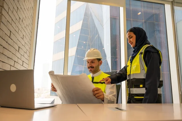 مهندسان معماريان سعوديان خليجيان يحملان مخطط لمشروع البناء للتخطيط وتنفيذ العمل في داخل المكتب في المملكة العربية السعودية, إرتداء سترة الحماية الخاصة بالعمل, تدقيق العمل، الإشراف والنقاش في العمل، مفهوم الهندسة، مهن ووظائف هندسية، دمج التكنولوجيا في العمل