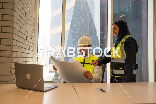الإشراف والنقاش في العمل، مهندسان معماريان سعوديان خليجيان يحملان مخطط لمشروع البناء للتخطيط وتنفيذ العمل في داخل المكتب في المملكة العربية السعودية, إرتداء سترة الحماية الخاصة بالعمل, تدقيق العمل،  مفهوم الهندسة، مهن ووظائف هندسية،دمج التكنولوجيا في العمل