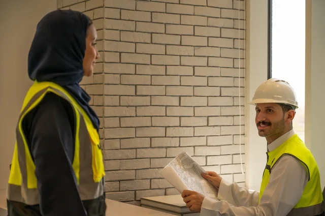 مهن ووظائف هندسية، الإشراف والنقاش في العمل، صورة مقربة لمهندسان معماريان سعوديان خليجيان يحملان مخطط لمشروع البناء للتخطيط وتنفيذ العمل في داخل المكتب في المملكة العربية السعودية, دمج التكنولوجيا في العمل، إرتداء سترة الحماية الخاصة بالعمل, تدقيق العمل