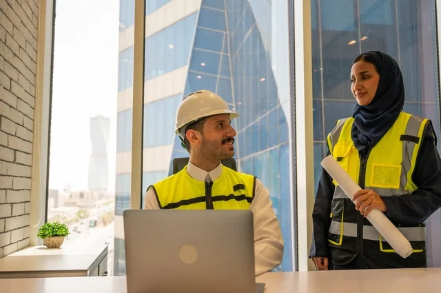 Writing and recording data and results, managing and completing tasks using modern and advanced technical devices, a profession and office job in Saudi Arabia, two Saudi Gulf architects carrying a plan for a construction project to plan and execute work inside the office, wearing a work safety vest, auditing the work.