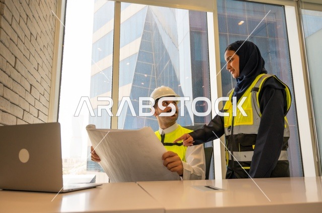 مهنة ووظيفة مكتبية سعودية، مهندسان معماريان سعوديان خليجيان يحملان مخطط لمشروع البناء للتخطيط وتنفيذ العمل في داخل المكتب, إرتداء سترة الحماية الخاصة بالعمل, تدقيق العمل،  كتابة وتدوين البيانات والنتائج، ادارة وانجاز المهام باستخدام جهاز تقني حديث ومتطور