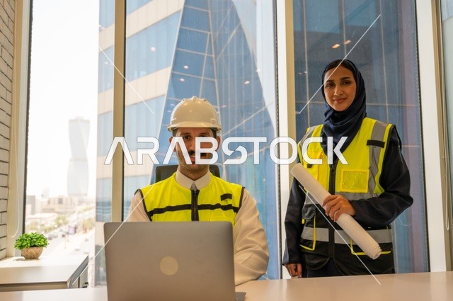 Managing and completing tasks using a modern and advanced technical device, a profession and office job in Saudi Arabia, two Saudi Gulf architects carrying a plan for a construction project to plan and execute work inside the office, wearing work safety vests, reviewing work, writing and recording data and results.
