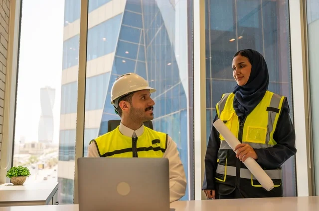 Work auditing, managing and completing tasks using modern and advanced technical devices, a profession and office job in Saudi Arabia, two Saudi Gulf architects holding a plan for a construction project to plan and execute work inside the office, wearing work safety vests, writing and recording data and results.