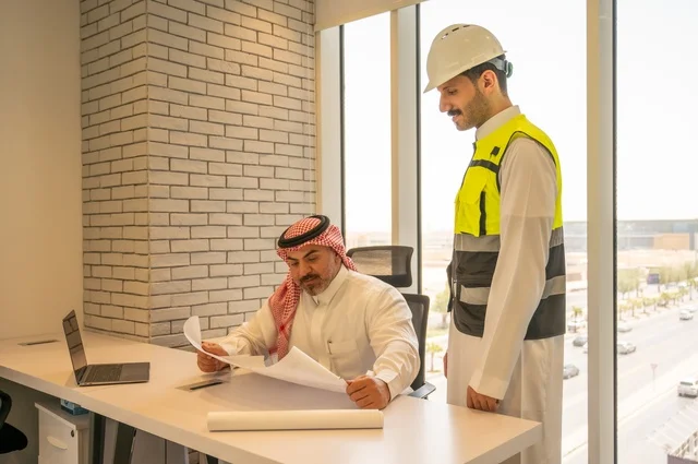 مفهوم الهندسة والإنشاءات المعمارية, مشاريع إعمارية سعودية وطنية، التفاهم والتعاون بين الزملاء، مهندس عربي خليجي سعودي يرتدي سترة العمل وخوذة الحماية البيضاء مع زميله يقرأوا المخططات الهندسية يقوموا بتدوين الملاحظات باستخدام الأجهزة التقنية الحديثة