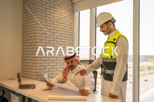 مشاريع إعمارية سعودية وطنية،التفاهم والتعاون بين الزملاء، مهندس عربي خليجي سعودي يرتدي سترة العمل وخوذة الحماية البيضاء يقف مع زميله يقرأوا المخططات الهندسية، متابعة مخطط المشروع الهندسي وتدقيقه, مفهوم الهندسة والإنشاءات المعمارية