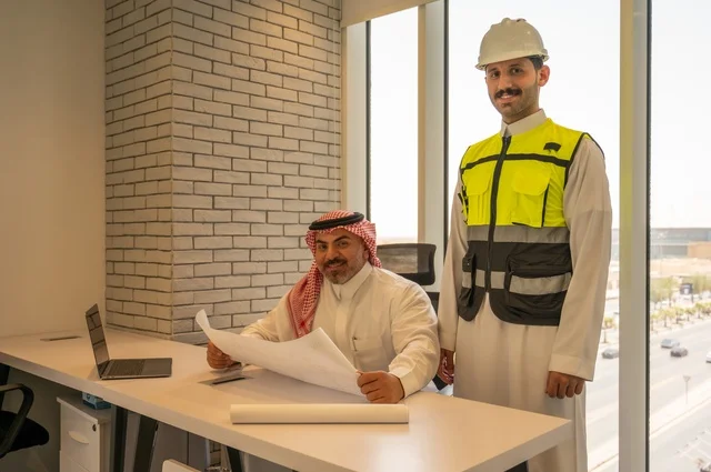 تبادل الخبرات والمعلومات لتحقيق الاهداف،،التفاهم والتعاون بين الزملاء، مهندس عربي خليجي سعودي يرتدي سترة العمل وخوذة الحماية البيضاء يقف مع زميله، متابعة مخطط المشروع الهندسي وتدقيقه, مفهوم الهندسة والإنشاءات المعمارية، مشاريع إعمارية سعودية وطنية