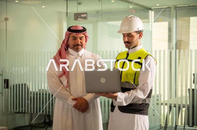 التفاهم والتعاون بين الزملاء، تبادل الخبرات والمعلومات لتحقيق الاهداف، مهندس عربي خليجي سعودي يرتدي سترة العمل وخوذة الحماية البيضاء يقف مع زميله، متابعة مخطط المشروع الهندسي وتدقيقه, مفهوم الهندسة والإنشاءات المعمارية، مشاريع إعمارية سعودية وطنية
