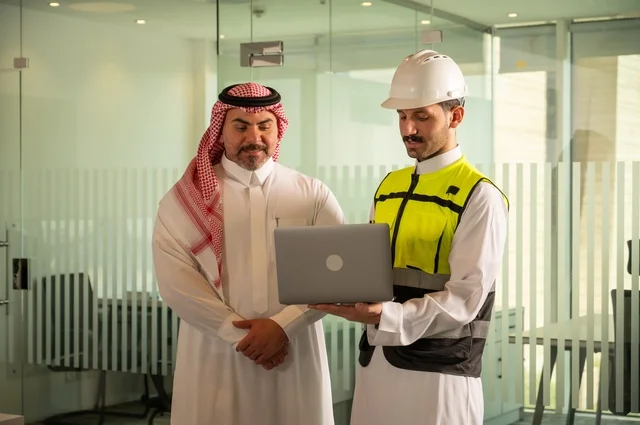 Understanding and cooperation among colleagues, exchanging experiences and information to achieve goals, a Saudi Gulf Arab engineer wearing a work vest and a white safety helmet stands with his colleague, following up on and reviewing the engineering project plan, the concept of engineering and architectural constructions, national Saudi construction projects.