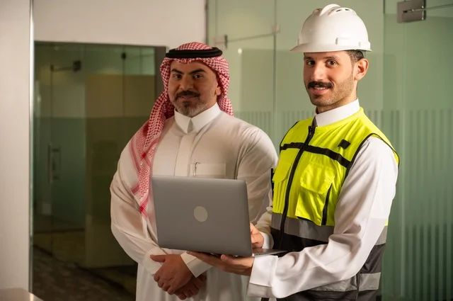 تبادل الخبرات والمعلومات لتحقيق الاهداف،،التفاهم والتعاون بين الزملاء، مهندس عربي خليجي سعودي يرتدي سترة العمل وخوذة الحماية البيضاء يقف مع زميله، متابعة مخطط المشروع الهندسي وتدقيقه, مفهوم الهندسة والإنشاءات المعمارية، مشاريع إعمارية سعودية وطنية