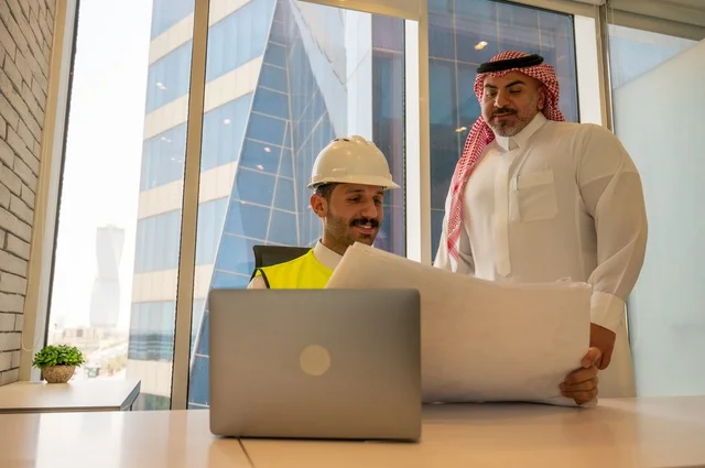 مهندس عربي خليجي سعودي يرتدي سترة العمل وخوذة الحماية البيضاء مع زميله يقرأوا المخططات الهندسية يقوموا بتدوين الملاحظات باستخدام الأجهزة التقنية الحديثة، التفاهم والتعاون بين الزملاء، مفهوم الهندسة والإنشاءات المعمارية, مشاريع إعمارية سعودية وطنية