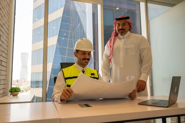 Gestures of happiness and joy, a meeting in the office, agreement and partnership on business and projects, signing new business deals, a side shot of a Saudi Arabian Gulf man wearing a thobe and ghutrah with his colleague, an engineer wearing a helmet and safety vest, national Saudi construction projects.