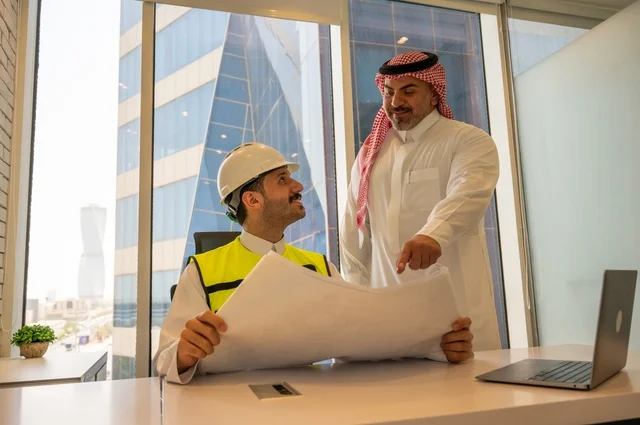 اجتماع في مكتب العمل، ايماءات السعادة والسرور،الاتفاق والشراكة على الأعمال والمشاريع، عقد صفقات عمل جديدة، تصوير من الجانب لرجل عربي خليجي سعودي يرتدي الثوب والشماغ مع زميله مهندس يرتدي الخوذة وسترة الحماية,  مشاريع إعمارية سعودية وطنية