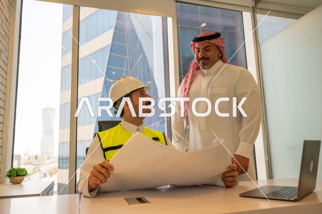 الاتفاق والشراكة على الأعمال والمشاريع، اجتماع في مكتب العمل، ايماءات السعادة والسرور،عقد صفقات عمل جديدة، تصوير من الجانب لرجل عربي خليجي سعودي يرتدي الثوب والشماغ مع زميله مهندس يرتدي الخوذة وسترة الحماية,  مشاريع إعمارية سعودية وطنية