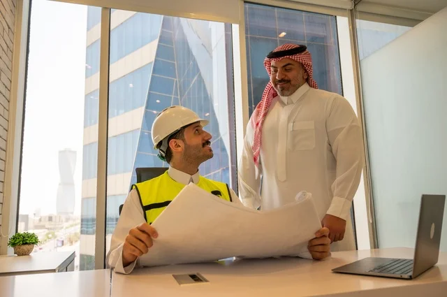 الاتفاق والشراكة على الأعمال والمشاريع، اجتماع في مكتب العمل، ايماءات السعادة والسرور،عقد صفقات عمل جديدة، تصوير من الجانب لرجل عربي خليجي سعودي يرتدي الثوب والشماغ مع زميله مهندس يرتدي الخوذة وسترة الحماية,  مشاريع إعمارية سعودية وطنية