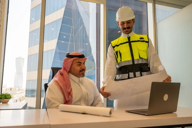 مشاريع إعمارية سعودية وطنية،التفاهم والتعاون بين الزملاء، مهندس عربي خليجي سعودي يرتدي سترة العمل وخوذة الحماية البيضاء يقف مع زميله يقرأوا المخططات الهندسية، متابعة مخطط المشروع الهندسي وتدقيقه, مفهوم الهندسة والإنشاءات المعمارية