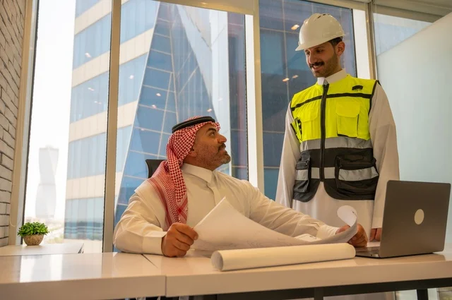 ايماءات السعادة والسرور، اجتماع في مكتب العمل، الاتفاق والشراكة على الأعمال والمشاريع، عقد صفقات عمل جديدة، تصوير من الجانب لرجل عربي خليجي سعودي يرتدي الثوب والشماغ مع زميله مهندس يرتدي الخوذة وسترة الحماية,  مشاريع إعمارية سعودية وطنية