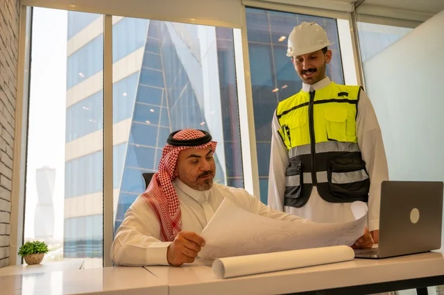 مهندس عربي خليجي سعودي يرتدي سترة العمل وخوذة الحماية البيضاء مع زميله يقرأوا المخططات الهندسية يقوموا بتدوين الملاحظات باستخدام الأجهزة التقنية الحديثة، التفاهم والتعاون بين الزملاء، مفهوم الهندسة والإنشاءات المعمارية, مشاريع إعمارية سعودية وطنية